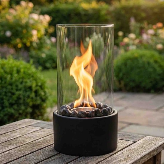 Tafelhaard met vlam in glazen cilinder, op zwarte basis met stenen, op een houten tafel in de tuin.