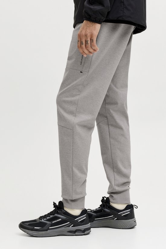 Detail van grijze joggingbroek met tekst 'JJ ATHLETIC DEPT.' op de zijkant. Zwarte sneakers.