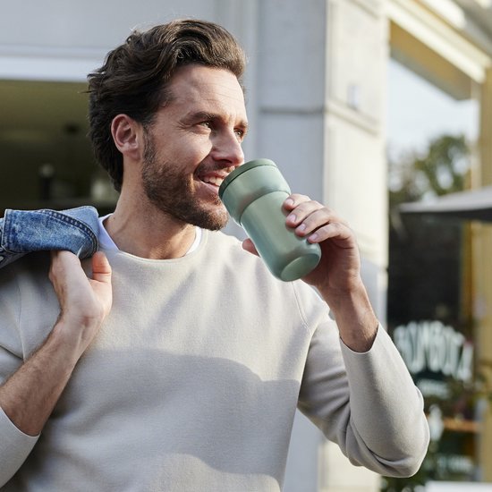 Homme buvant d'une tasse vert clair dans ses mains à l'extérieur.