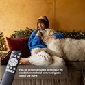 Vrouw met hond op bank, houdt afstandsbediening vast; past licht en ventilator aan.