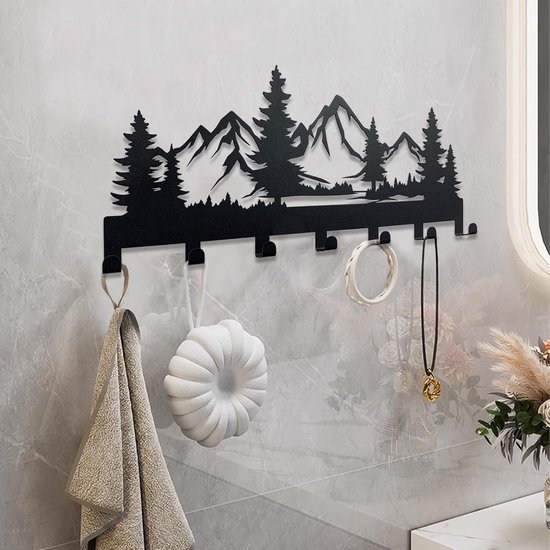 Zwart metalen sleutelhanger met bergen en bomen silhouet, hangt aan een muur met een handdoek, zeep en sieraden.