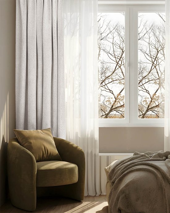 Lichtgrijs verduisterend gordijn naast een beige fauteuil en bed bij een raam met kale takken.