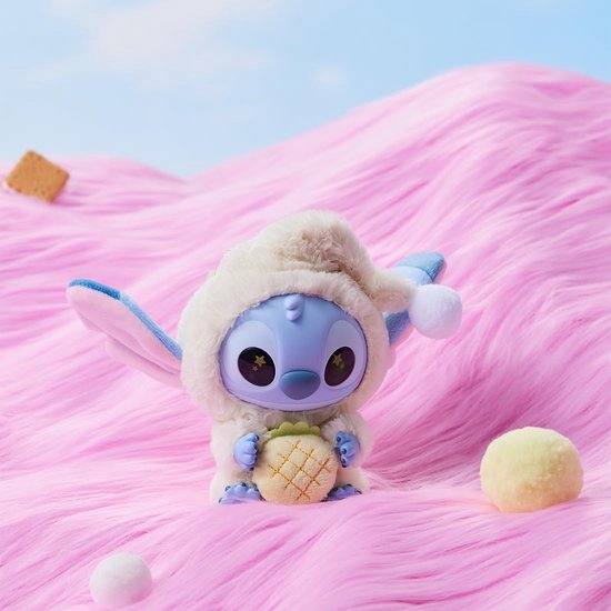 Een Stitch pluche speelgoedje in een crème slaapmutsje, met een ananas in de handen, op roze heuvels.