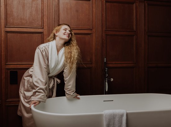 Vrouw in satijnen badjas lacht in een badkamer met houten lambrisering.