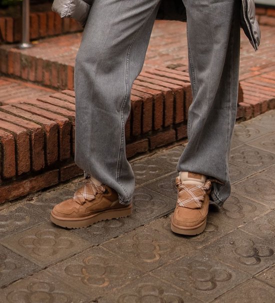 Model draagt bruine UGG sneakers met ruitpatroon veters op een betegeld terras.