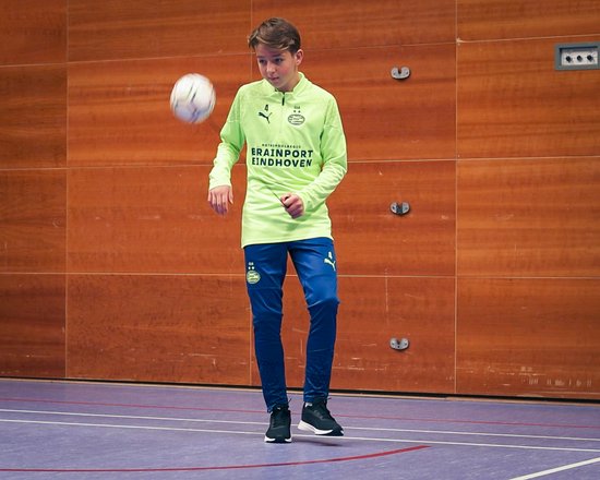 Jongen in een fluogroen sportshirt met lange mouwen, blauwe voetbalbroek en zwarte schoenen, trapt een bal in een sporthal.