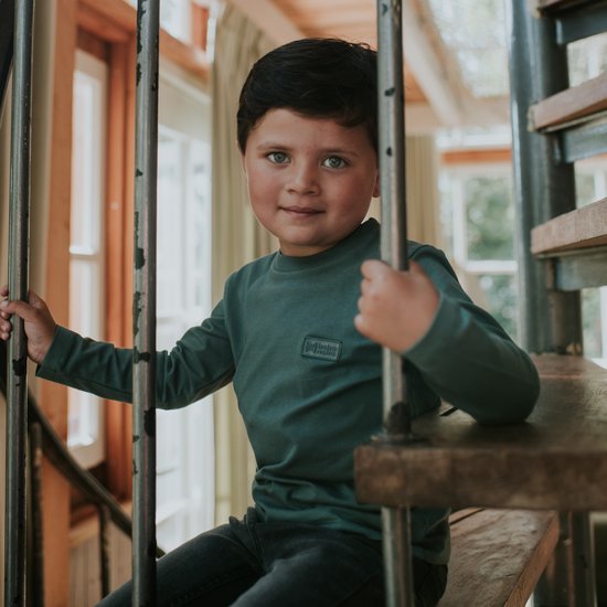 Jongen met donkergroen T-shirt met lange mouwen, zit op een houten trap.