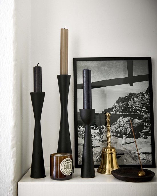 Drie zwarte, slanke kandelaren met verschillende hoogtes, met zwarte kaarsen, naast een bronzen kandelaar met een gouden bel en een brandende wierookstok op een marmeren oppervlak.