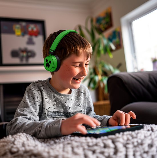 Jongen met een glimlach draagt een groene Minecraft koptelefoon met Creeper-print en kijkt naar een tablet.