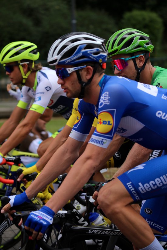 Wielrenners in blauw-witte, witte en groene tenues, dragen helmen en zonnebrillen.