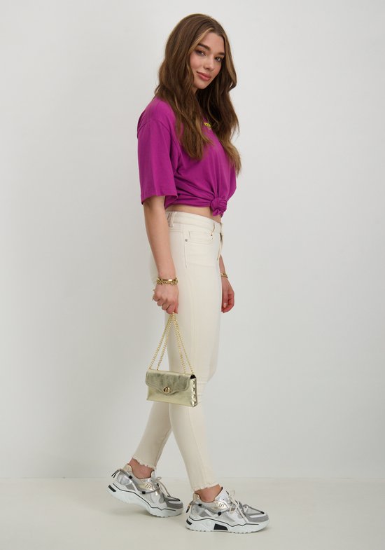 Model met een paarse top, beige skinny jeans, gouden tasje en zilverkleurige sneakers.