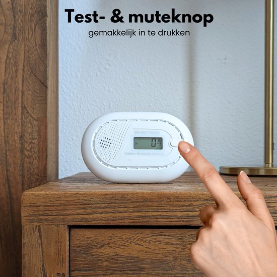 Vinger drukt op de testknop van een witte koolmonoxidemelder op een nachtkastje.
