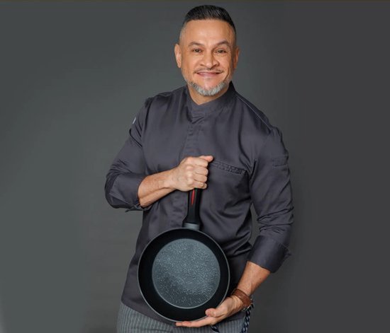Homme d'âge moyen avec une barbe grise, vêtu d'une veste de chef grise, tenant une poêle wok noire à revêtement marbré.