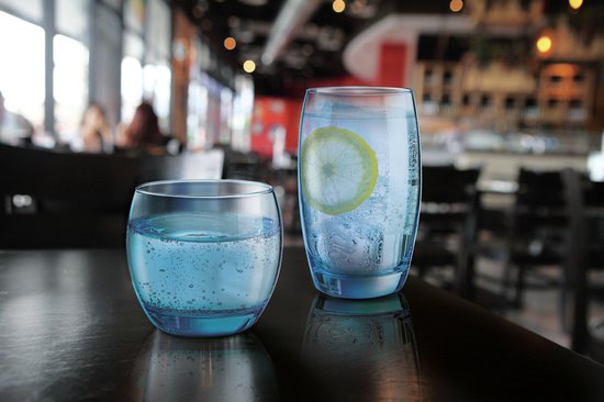 Twee glazen op een tafel: een met bruisend water, ijs en citroen, de ander met blauw water.