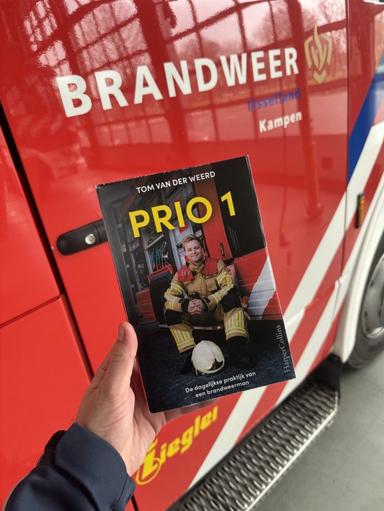 Een hand houdt het boek "PRIO 1" vast, met de omslag zichtbaar, bovenop een brandweerwagen.