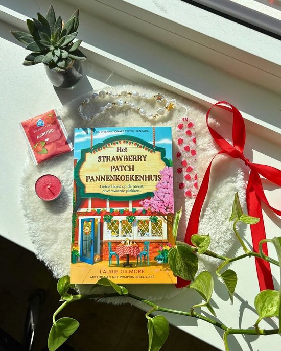Livre "Het Strawberry Patch Pannenkoekenhuis" sur un tapis blanc et moelleux, avec des plantes, une bougie, du thé et un marque-page aux fraises.