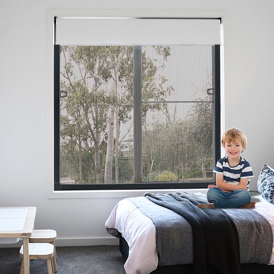 Een jongetje zit op een bed in een slaapkamer met uitzicht op bomen door een horraam.