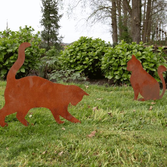 Twee roestkleurige metalen katten silhouetten in een grasveld, met struiken op de achtergrond.