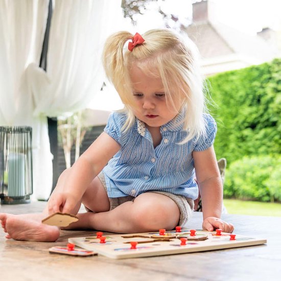 Een jong meisje met blonde paardenstaart zit op een tafel en speelt met een houten puzzel.