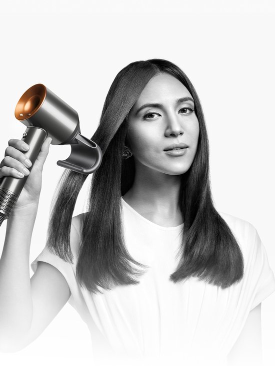 Vrouw met lang steil haar, gebruikt een zilverkleurige Dyson föhn met koperkleurige details.