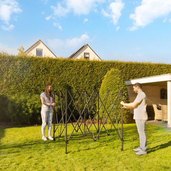 Een man en een vrouw zetten een zwart partytent frame op in een grasveld met een heg en huizen op de achtergrond.
