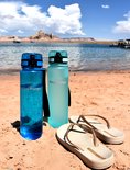Kleine blauwe en lichtblauwe King Mungo drinkflessen op een strand met teenslippers.