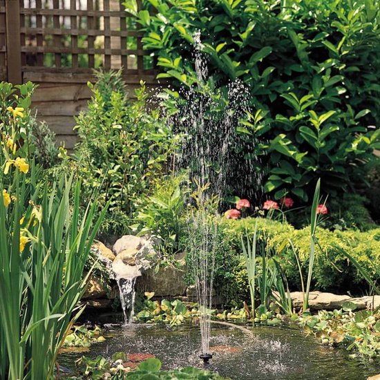 Een fontein spuit water in een vijver met groene waterplanten en een houten schutting.