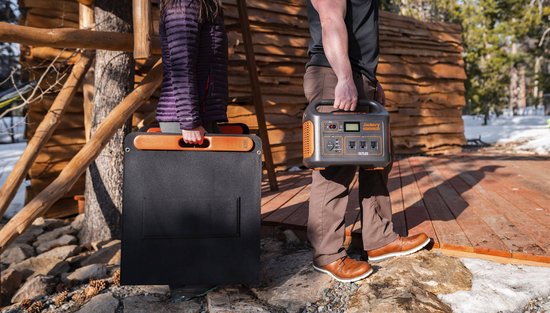 Man met Jackery Explorer 1500 en vrouw met Jackery SolarSaga 100, beide in buitenseizoen kleding, bij houten hut in de sneeuw.