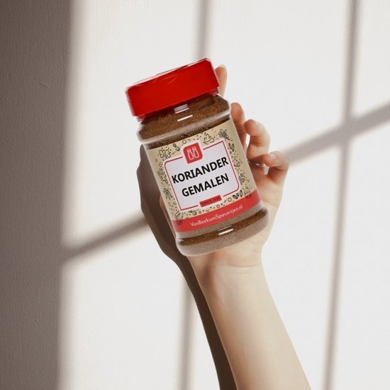 Een hand houdt een plastic flesje met gemalen koriander vast. Het flesje heeft een rood deksel en een beige etiket met de tekst 'KORIANDER GEMALEN'.