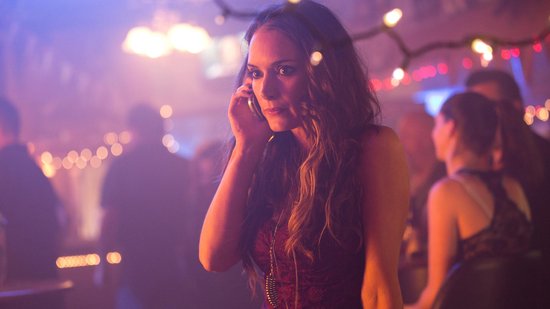 Een vrouw met lang bruin haar, gekleed in een rode kanten jurk, praat aan de telefoon in een schemerige bar.