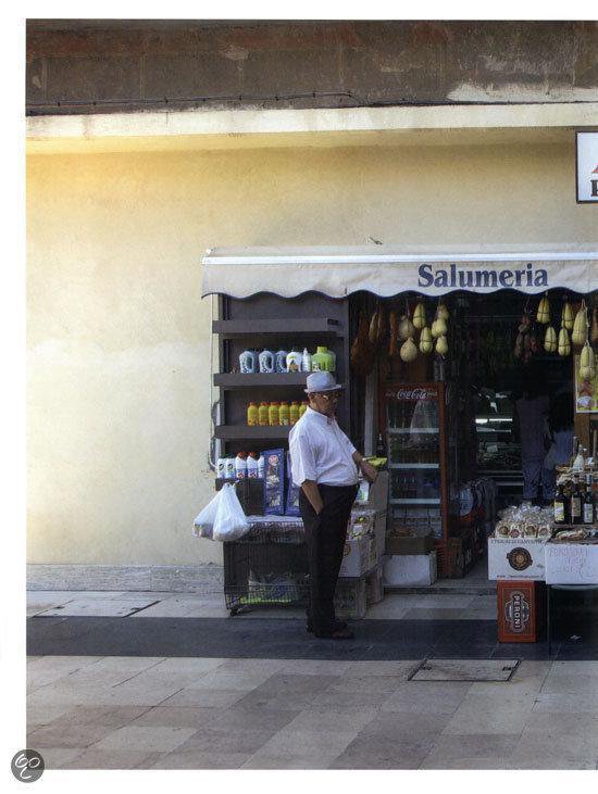 Man in wit shirt en hoed bij een winkel met de tekst 'Salumeria'.