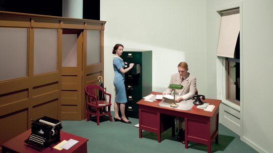 Een vrouw in een blauwe jurk staat bij een archiefkast, een man zit aan een bureau.