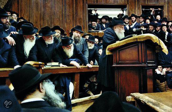 Groep orthodoxe Joden, mannen met baarden en hoeden, luisteren naar een spreker bij een houten katheder.