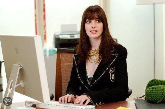 Vrouw met pony, goudkleurige ketting en zwart jasje met zilveren details, zittend achter een bureau.