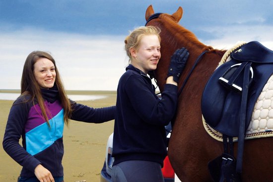 Twee jonge vrouwen met een bruin paard op een strand. Vrouw links met paars-blauwe jas, vrouw rechts met zwarte jas.