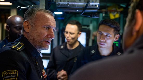 Man in donker marine-uniform met Russische tekst op de mouw spreekt met andere officieren
