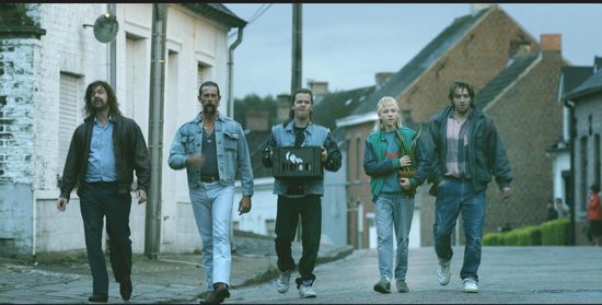 Vijf mannen lopen in een straat, gekleed in jaren 80 kleding. Eén man draagt een spijkerjasje.