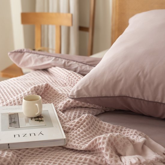 Een close-up van een lila kussen met een boek en een kopje koffie op een lila wafelsprei.