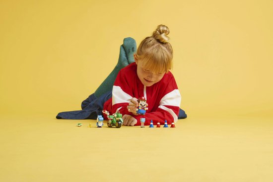 Blond meisje in rode jas ligt op gele achtergrond, speelt met LEGO Yoshi-Crosser.