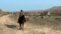 Man op een zwart paard rijdt op een stoffige weg, met een klein huis en cactussen op de achtergrond.