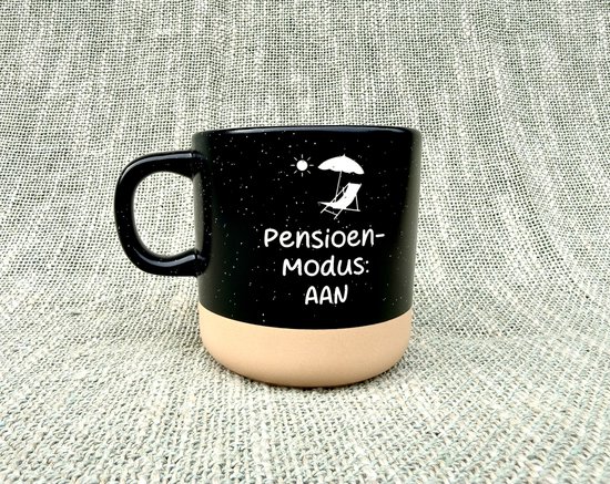 Zwarte mok met witte spikkels, beige onderkant en de tekst "Pensioen- Modus: AAN" met een zon en ligstoel.