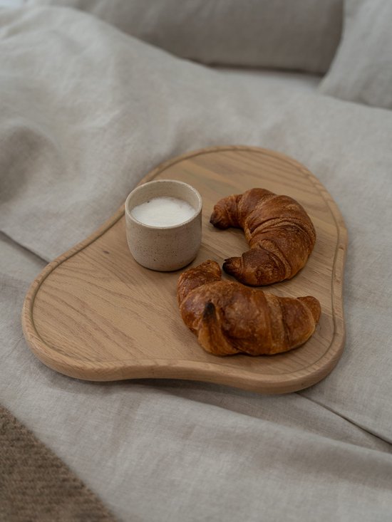 Houten dienblad met twee croissants en een kopje melk op een beige linnen bed.