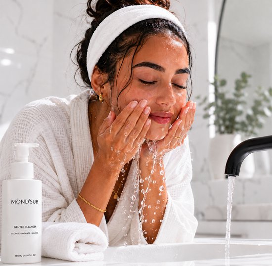 Een vrouw met een witte haarband wast haar gezicht in een luxe badkamer met een witte fles Mond'sub Gentle Cleanser ernaast.