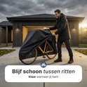 Man in donkere jas onthult gele fiets onder zwarte hoes op oprit.