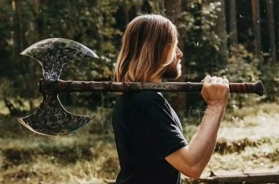 Man met lang blond haar draagt een dubbelbladige bijl met grijs, verweerd blad over zijn schouder.