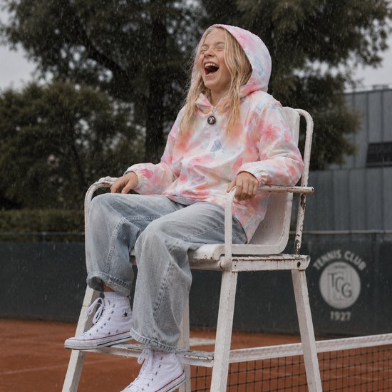 Lachend meisje in een regenjas met bloemenprint in pastelkleuren, zittend op een tennisscheidsrechtersstoel.