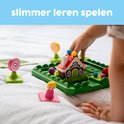 Hand van een kind dat de stukjes van het spel Hans & Grietje plaatst. Speelgoedhuisje, personages, ballen, snoep en speelbord zichtbaar.