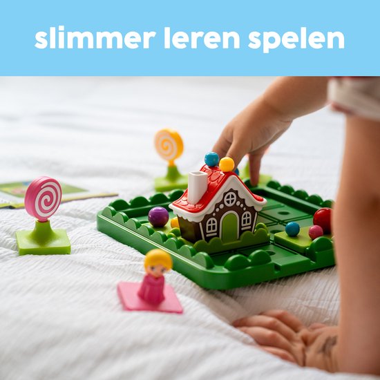 Hand van een kind dat de stukjes van het spel Hans & Grietje plaatst. Speelgoedhuisje, personages, ballen, snoep en speelbord zichtbaar.
