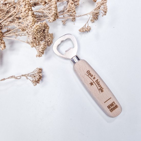 Houten flesopener met 'Opa's biertje - Geniet ervan!' en ster. Neutrale achtergrond met gedroogde bloemen.