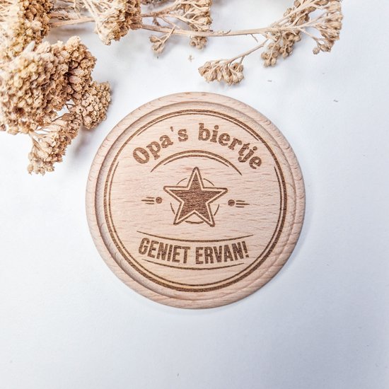 Houten onderzetter met 'Opa's biertje - Geniet ervan!' en ster. Neutrale achtergrond met gedroogde bloemen.